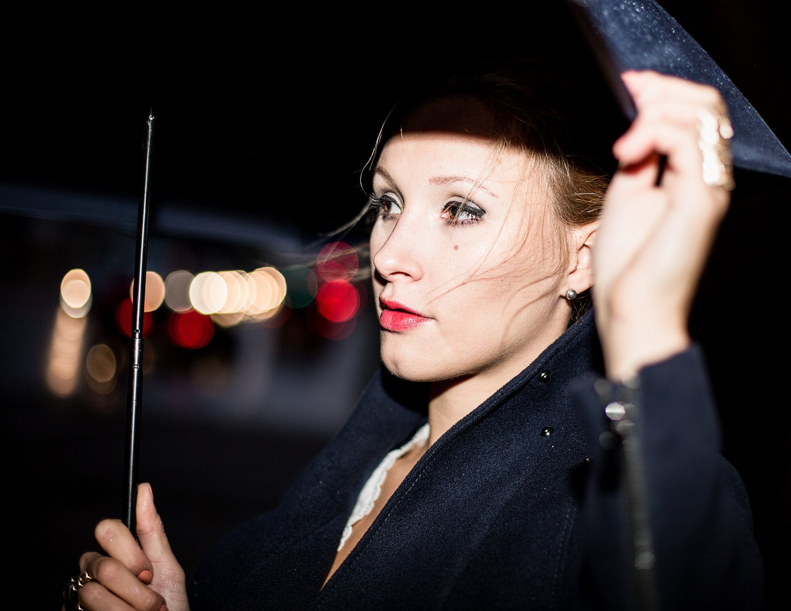 Eine Frau unter einem Regenschirm, Close-up, bei Nacht, verschwommene Lichter einer befahrenen Straße im Hintergrund.