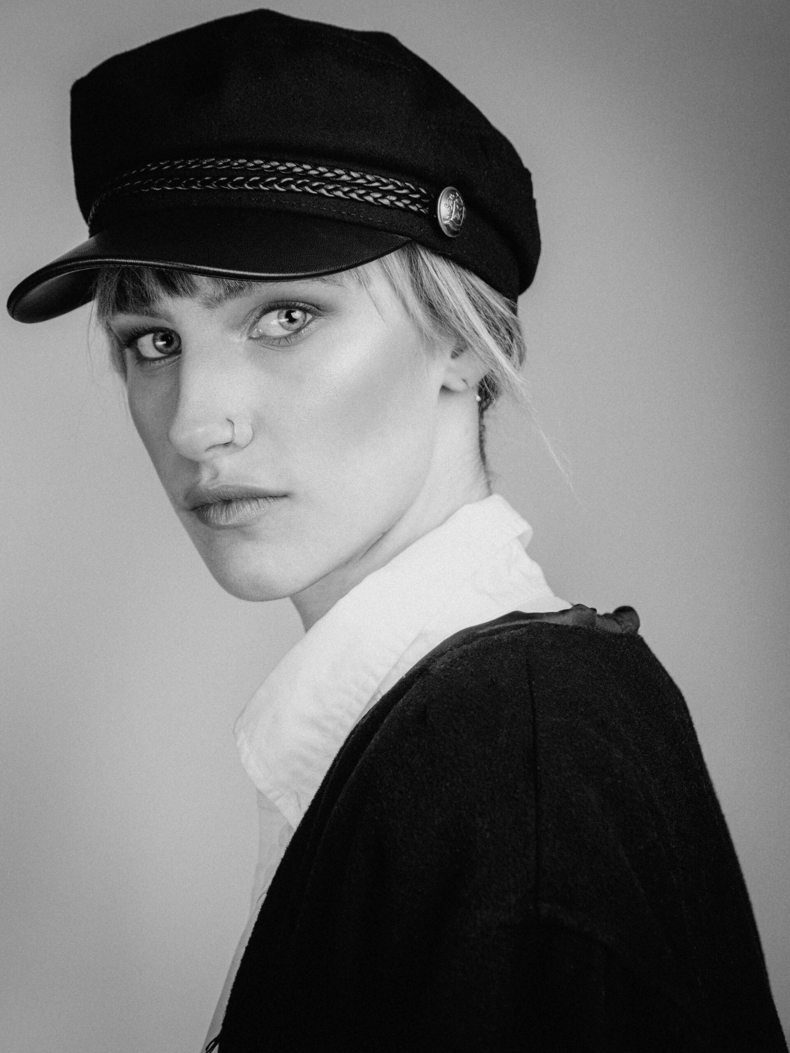 Studio Porträt einer Frau, die eine Kappe trägt. Sie steht seitlich und dreht den Kopf zum Betrachter. Schwarzweiss Fotografie