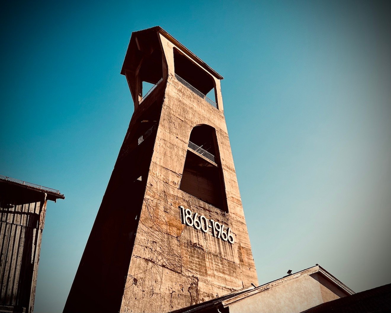 Froschperspektive des historischen Förderturms des ehemaligen Bergwerks Hausham, aufgenommen vor klarem blauem Himmel. An der Betonfassade prangt in weißen Ziffern „1860–1966" – die Betriebsjahre des Kohlebergwerks. Der Turm verjüngt sich nach oben und öffnet sich in zwei große Bogenfenster sowie eine offene Plattform mit Überdachung an der Spitze. Vertrocknete Rankpflanzen ziehen sich über den verwitterten Beton. Links im Bild eine angrenzende Holzscheune. Die warme Lichtstimmung und leichte Vignettierung verleihen dem Industriedenkmal eine monumentale, fast ehrfürchtige Wirkung.