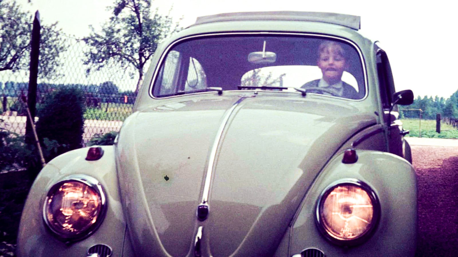 Vintage-Farbfotografie aus den 1972, mit typischem warmen Farbstich. Raimund sitzt lächelnd hinter dem Lenkrad eines beigefarbenen VW Käfers und schaut durch die Windschutzscheibe. Das Auto ist frontal aufgenommen, die runden Scheinwerfer und die geschwungene Motorhaube dominieren das Bild. Im Hintergrund ein Garten mit Maschendrahtzaun, Obstbäumen und Hecken – eine ländliche, sommerliche Szenerie.