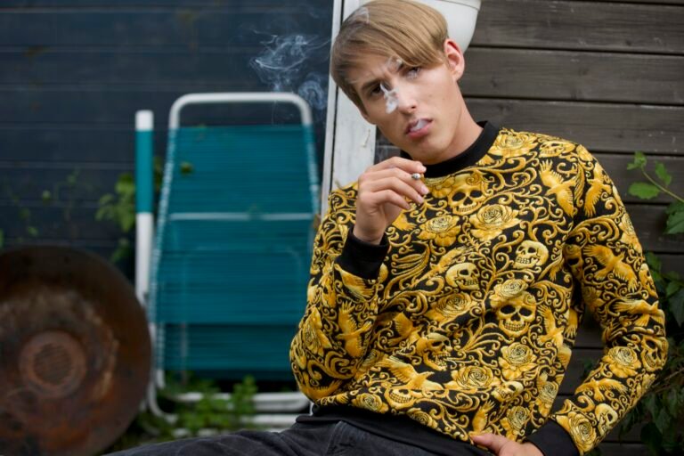 Photography Newsletter: A young man with light-colored hair leans against a weathered wooden wall. He is wearing a striking black and gold sweater with a skull and rose pattern and is holding a cigarette, the smoke curling in front of his face. His gaze is direct, slightly skeptical. Portrait session Raimund Verspohl (Rayhen) Photography, 2019   